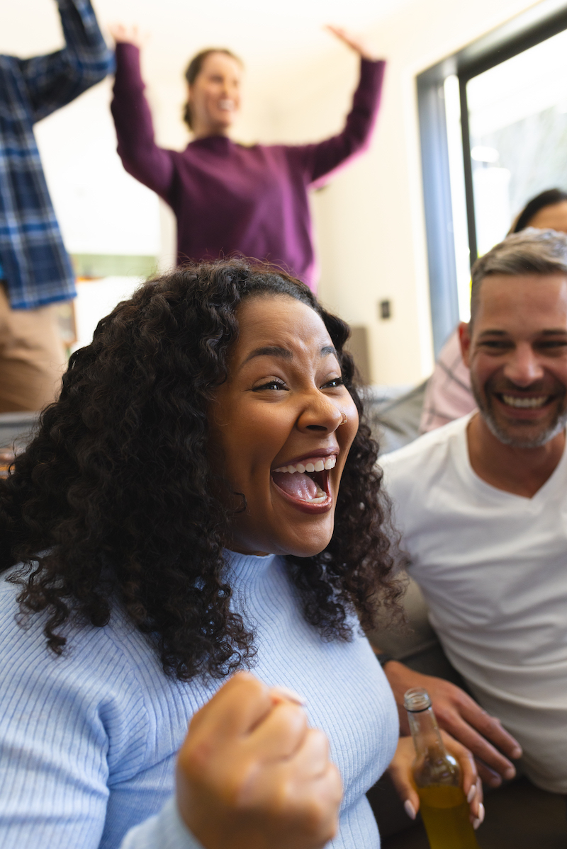 Excited diverse male and female friends watching sport on tv at home and drinking beers, copy space. Lifestyle, sport, competition, entertainment, friendship and domestic life, unaltered.